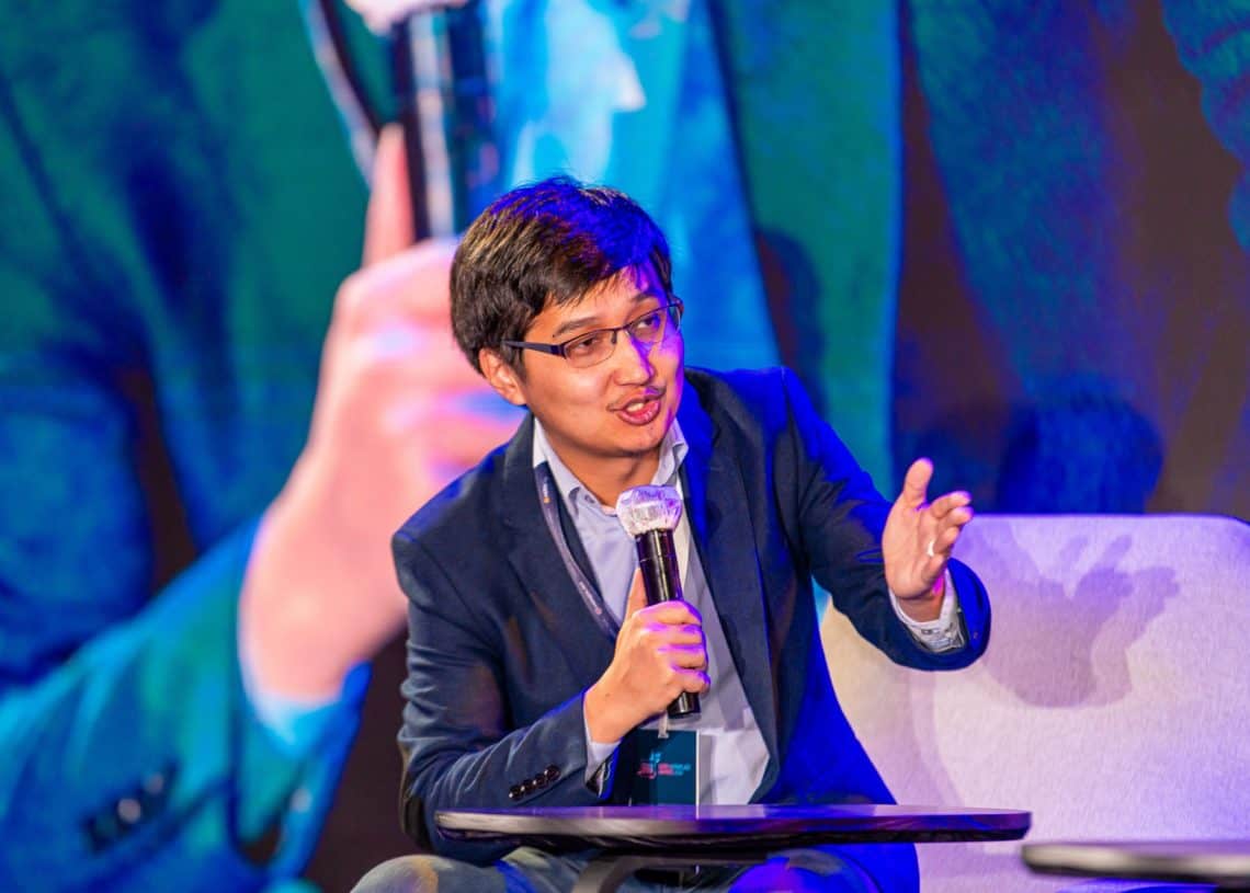 Монголын хөдөлмөрийн зах зээлийн дээд хэмжээний уулзалт 2024 дээр цэнхэр костюмтай хүн гараараа дохиж микрофонд ярьж байна. Тэд тайзан дээр сууж, том дэлгэцэн дээр өөрсдийн дүр төрхийг харуулахын тулд оролцогчдод хандаж, хувьсан өөрчлөгдөж буй хүний нөөцийн ландшафт дахь сорилтуудын шийдлийг хайж байна. - Business.MN