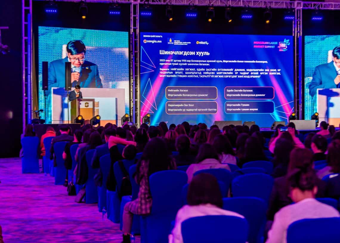 Монголын хөдөлмөрийн зах зээлийн дээд хэмжээний уулзалтын индэрт илтгэгч цэнхэр сандал дээр суусан үзэгчдэд хандаж үг хэлж байна. Шийдэл эрэлхийлэхийн хэрээр ард талын том дэлгэц нь текст болон график дүрслэлийг харуулж, өрөөг нил ягаан, цэнхэр өнгийн гэрлээр гэрэлтүүлдэг. - Business.MN