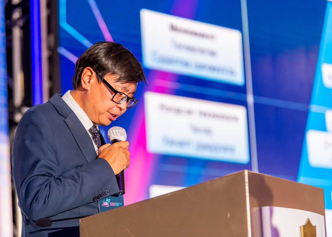 Костюм өмссөн хүн зогсож буй индэр дээр микрофоноор ярьж байна Монголын Хөдөлмөрийн зах зээлийн дээд хэмжээний уулзалт 2024. Арын дэвсгэр дээрх том дэлгэц нь хүний нөөцийн талаарх энэхүү чухал хурлын тайзыг текст болон графикаар харуулж байна. - Business.MN