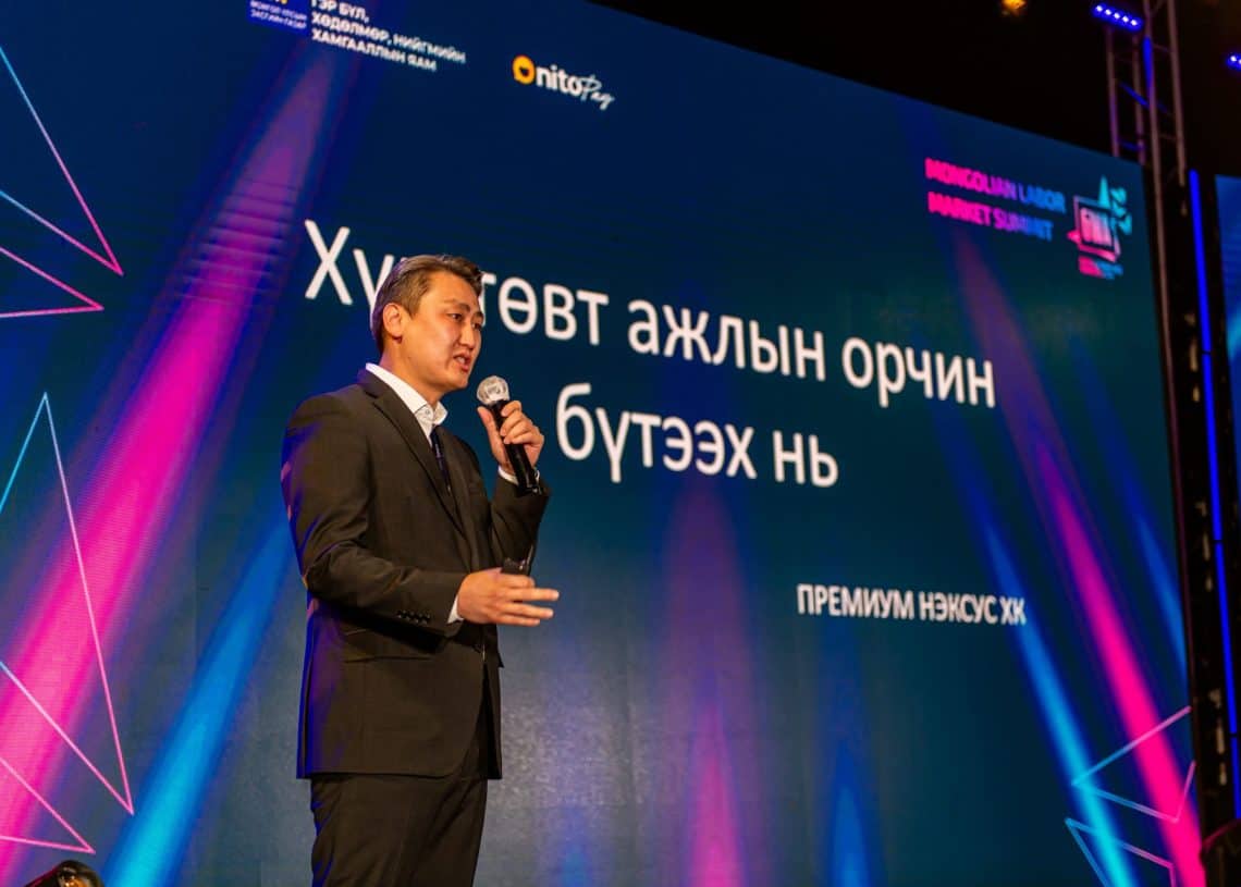 "Монголын хөдөлмөрийн зах зээлийн чуулга уулзалт-2024"-ийн тайзан дээр микрофон бариад "шийдэл хайсан" гэж нэг хүн үг хэлж байна. Тэдний ард байрлах том дэлгэц нь хөх, ягаан, нил ягаан өнгийн өнгөлөг геометрийн загвар бүхий монгол хэл дээрх бичвэрийг харуулдаг. - Business.MN