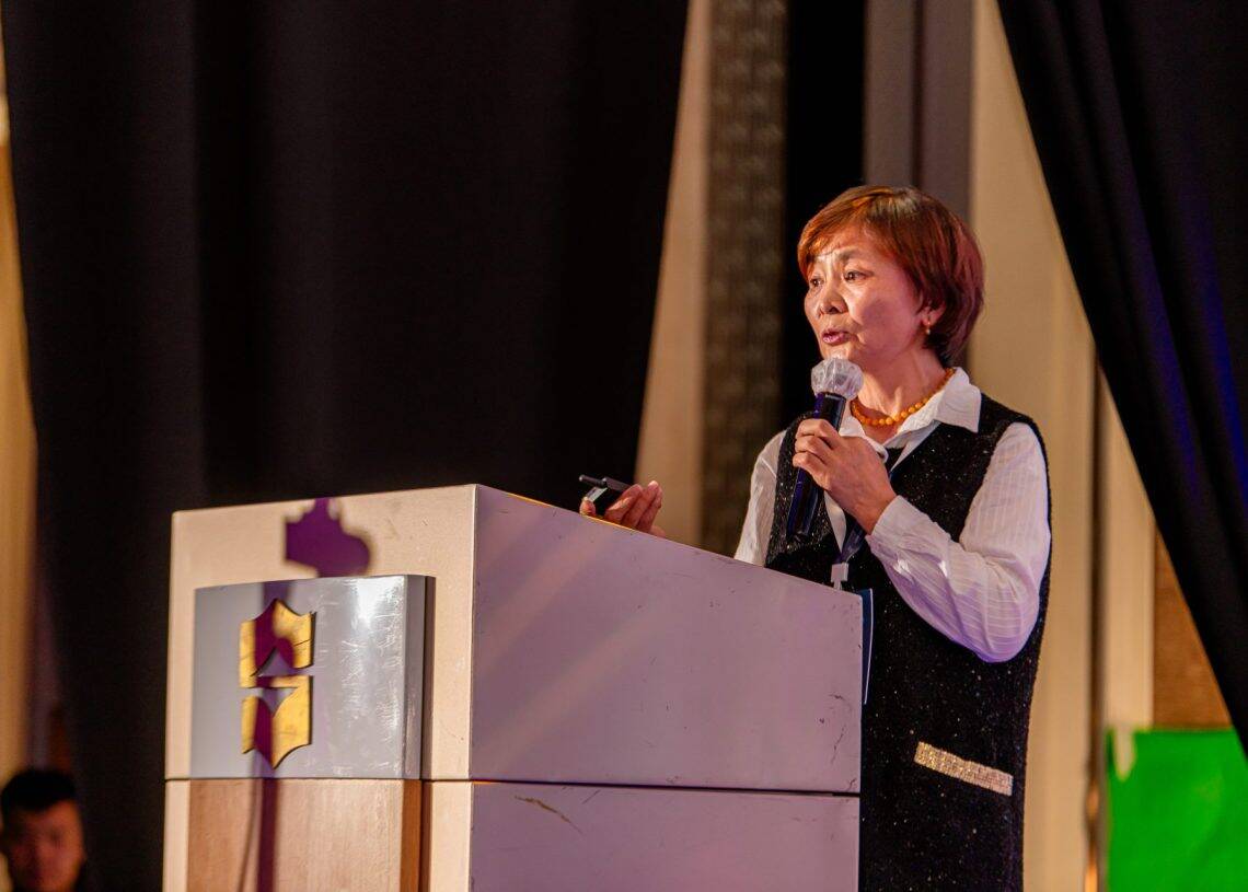 Монголын хөдөлмөрийн зах зээлийн дээд хэмжээний чуулга уулзалтыг сурталчилсан эмблем бүхий индэр дээр хүн микрофон барьж ярьж байна. Тэд богино үстэй, цагаан цамцан дээр хар хантааз өмсдөг. Харанхуй хөшиг, дулаан гэрэлтүүлэг нь хүний нөөц дэх шийдлийг хэлэлцэхэд тохиромжтой албан ёсны уур амьсгалыг бүрдүүлдэг. - Business.MN