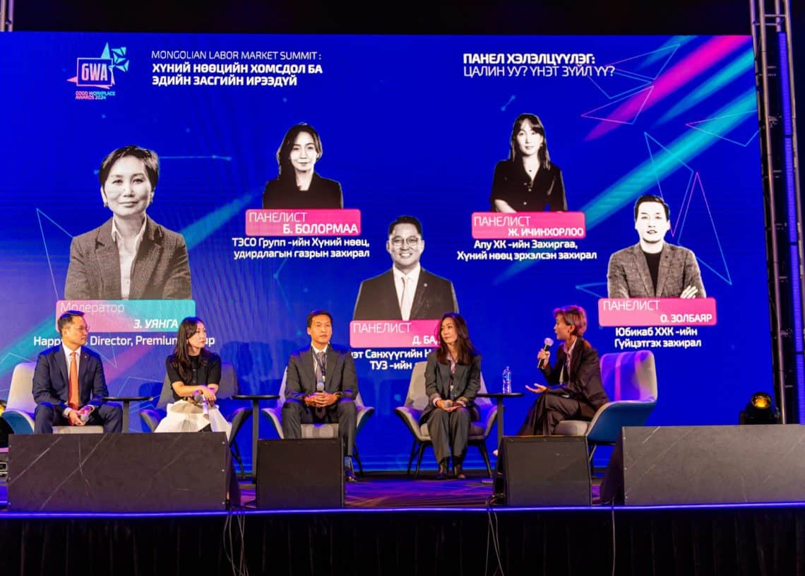 “Монголын хөдөлмөрийн зах зээлийн чуулга уулзалт-2024” уулзалтын тайзан дээр таван хүний бүрэлдэхүүнтэй хэсэг оролцогчид сууж байна. Тэдний ард том дэлгэцээр тэдний нэр, албан тушаал харагдана. Нил ягаан, цэнхэр өнгийн гэрэлтүүлгээр сайжруулсан албан ёсны орчин нь хүний нөөцийг судалж, тулгамдсан асуудлыг шийдвэрлэх арга замыг санал болгож буй өнгө аясыг бүрдүүлдэг. - Business.MN