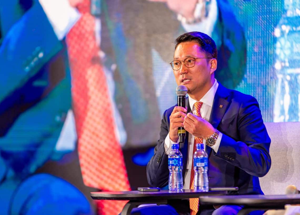 Цэнхэр костюмтай, нүдний шилтэй хүн Монголын хөдөлмөрийн зах зээлийн дээд хэмжээний уулзалтын үеэр тайзан дээр үзэгчдэд хандаж, хэлэлцүүлгийн үеэр санаа бодлоо илэрхийлж байна. Тэдний ард байгаа том дэлгэцэн дээр өөр чанга яригчийн бүдгэрсэн дүрс харагдах бөгөөд хоёр савтай усны сав ширээний ард тод харагдаж байна. - Business.MN