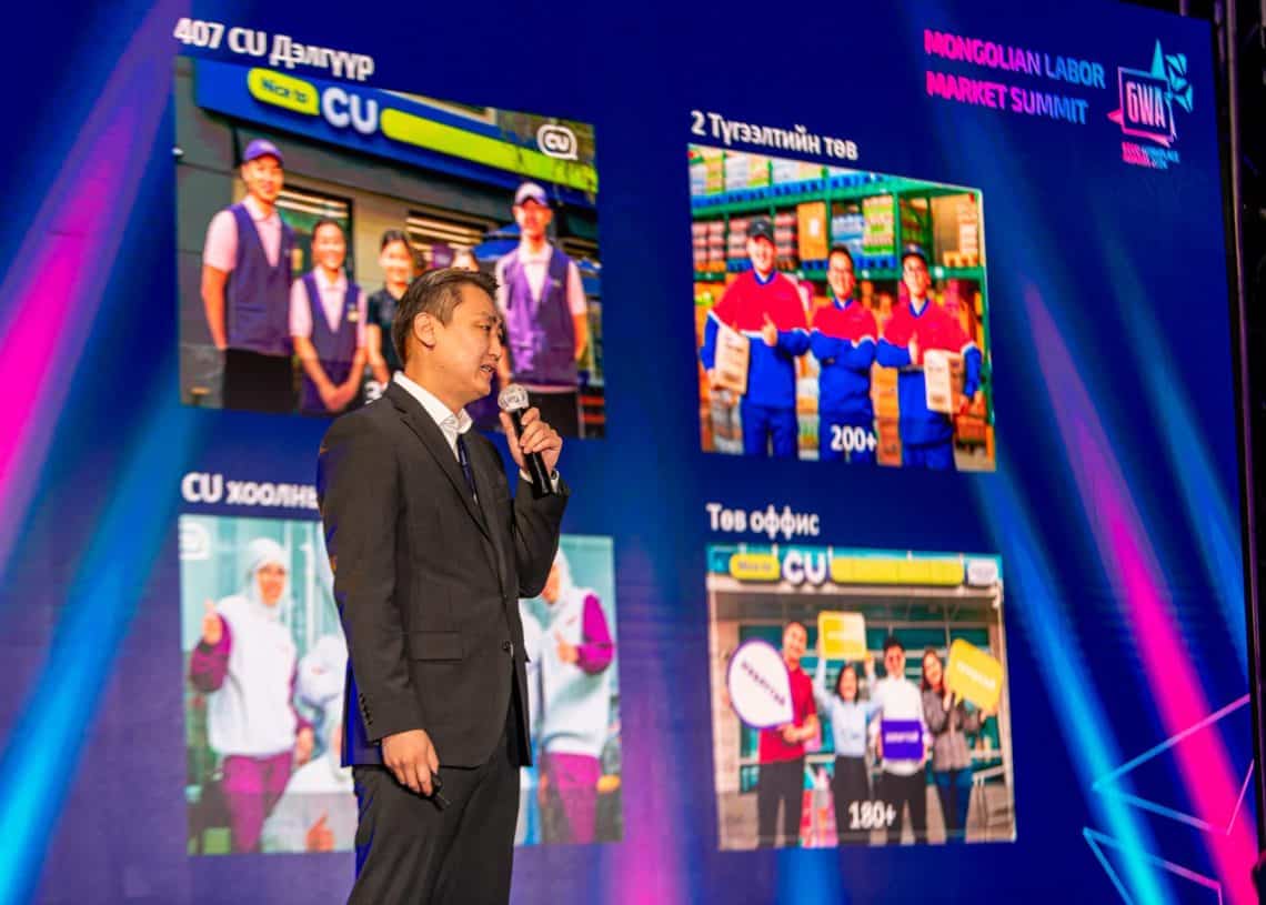 2024 оны Монголын хөдөлмөрийн зах зээлийн дээд хэмжээний чуулга уулзалтад илтгэгч оролцож, дүрэмт хувцас өмссөн ажилчдын зураг, "ЦУ" гэсэн бичээс бүхий том дэлгэцийн өмнө зогсож байна. Дэлгэцэнд хүний нөөцийн статистик мэдээлэл, CU-ын янз бүрийн байршлын гэрэл зургууд багтсан болно. - Business.MN