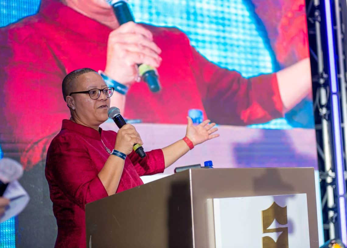 2024 оны Монголын хөдөлмөрийн зах зээлийн дээд хэмжээний уулзалтын үеэр улаан цамцтай хүн индэр дээр зогсож, микрофон барин урам зоригтойгоор дохиж байна. Дэлгэц дээрх тэдний томруулсан дүрс нь хүний нөөцийн талаарх гол санааг онцолж, энэхүү илтгэлийн арга хэмжээнд үзэгчдийг татан оролцуулж байна. - Business.MN