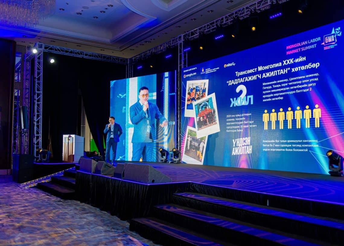 Костюм өмссөн илтгэгч тайзан дээр микрофон барин зогсож, 2024 оны дээд хэмжээний уулзалтын үеэр сонсогчдод хандан үг хэлж байна. Түүний арын том дэлгэц нь Монголын хөдөлмөрийн зах зээлийн тухай бичвэр, зураг, графикийг харуулдаг. Тохируулга нь албан ёсны бөгөөд цэнхэр, нил ягаан өнгийн гэрэлтүүлгээр сайжруулсан. - Business.MN