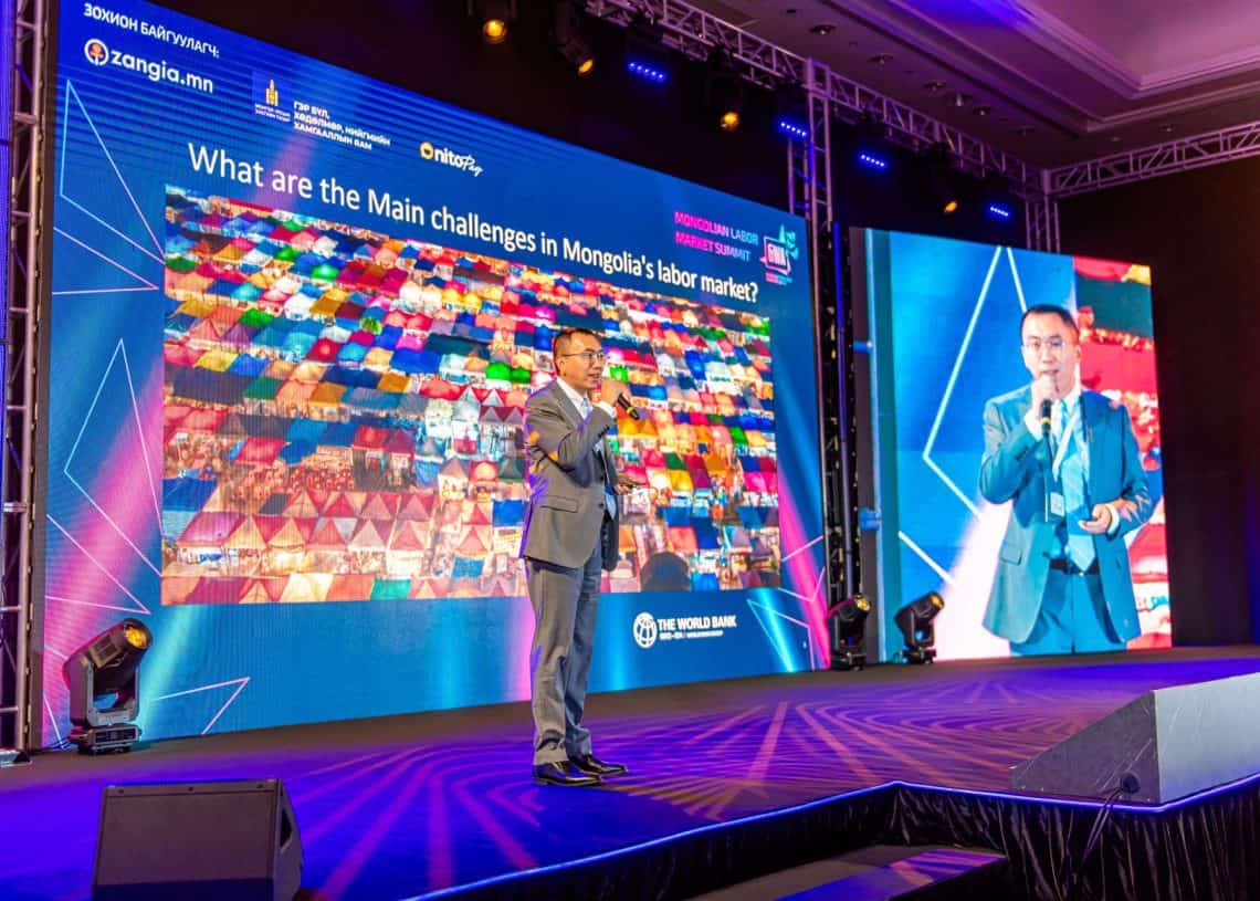 2024 оны Монголын хөдөлмөрийн зах зээлийн дээд хэмжээний уулзалтын үеэр костюм өмссөн илтгэгч тайзан дээр микрофон барин зогсоод хүний нөөцийн сорилтыг илтгэж байна. Түүний ард байрлах том дэлгэц дээр “Монголын хөдөлмөрийн зах зээлд тулгарч буй гол бэрхшээл юу вэ? - Business.MN