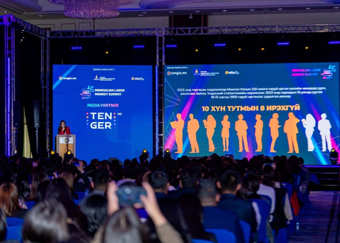 Монголын хөдөлмөрийн зах зээлийн дээд хэмжээний уулзалтын тайзны өөдөөс олон үзэгчид сууж байна. Зүүн талын индэр дээр хүн зогсож байна. Хурц гэрэлтдэг дэлгэц нь лааны суурь бүхий гоёл чимэглэлийн таазны доор шинэлэг шийдлийн талаар ярилцаж буй хүмүүсийн дүрсийг гадаад хэл дээрх бичвэрийн хажууд харуулж байна. - Business.MN