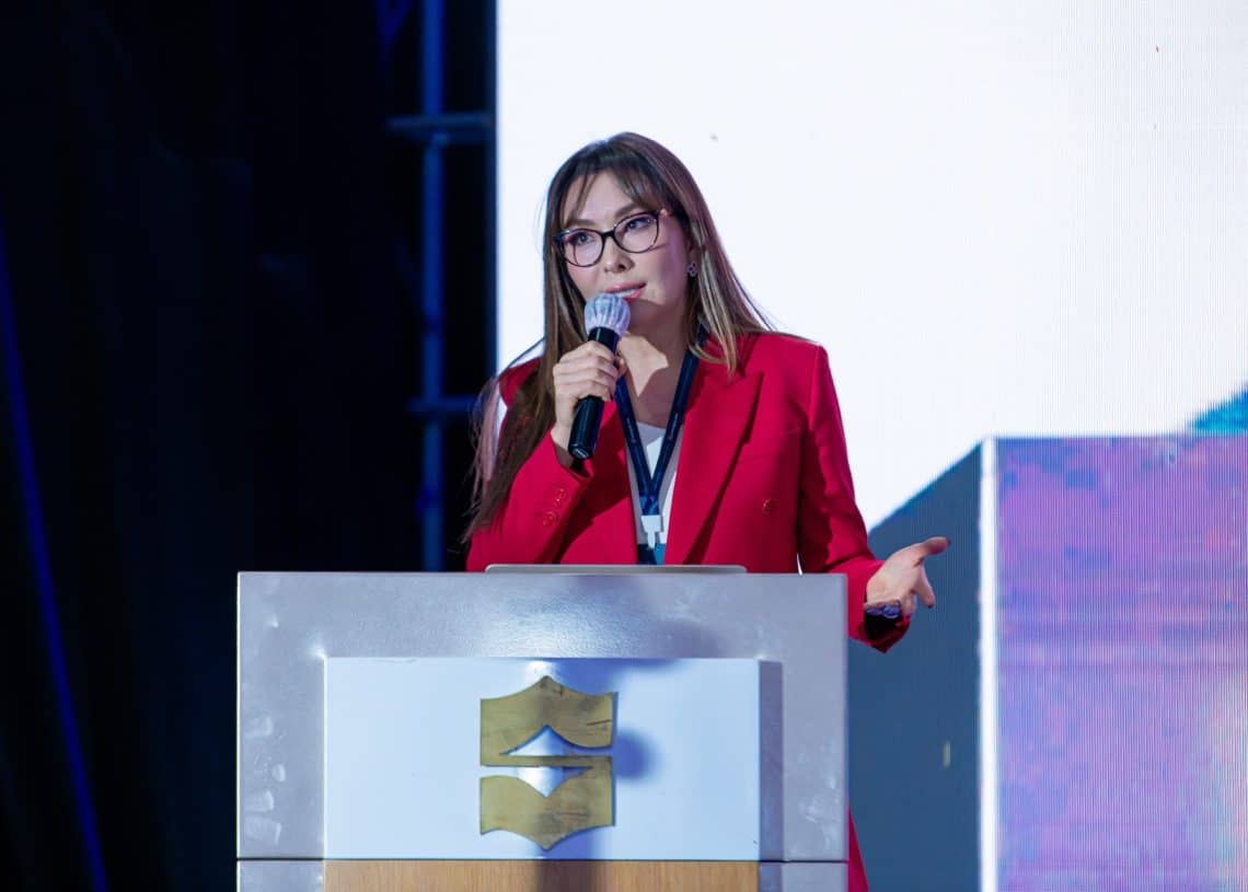 Улаан пиджак өмссөн хүн Монголын хөдөлмөрийн зах зээлийн чуулга уулзалт 2024 дээр бамбай тэмдгээр чимэглэсэн индэр дээр микрофоноор үг хэлж байна. Арын дэвсгэрийг хэсэгчлэн том дэлгэцээр гэрэлтүүлж, хүний нөөцийн талаар ярилцаж, асуудлын шийдлийг санал болгож байна. - Business.MN