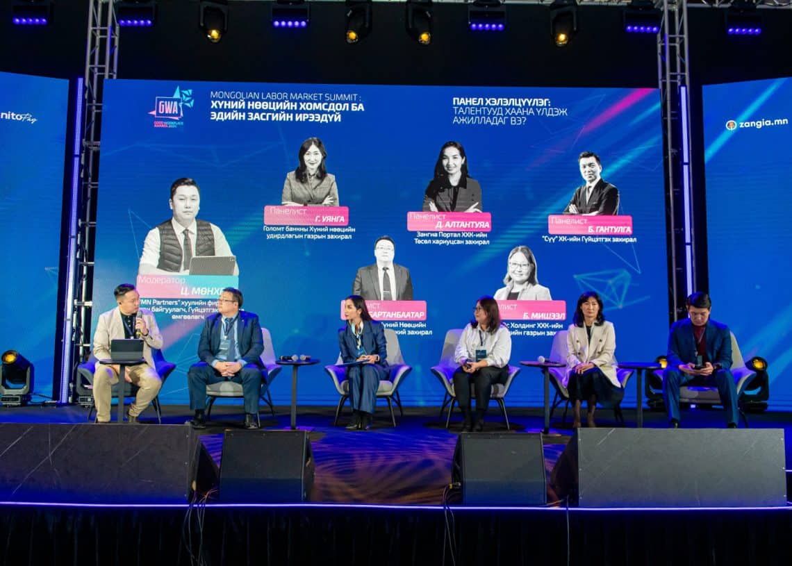 Монголын хөдөлмөрийн зах зээлийн дээд хэмжээний уулзалтын үеэр тайзан дээр суусан зургаан илтгэгчтэй зөвлөл үзэгчидтэй харилцаж байна. Тэдний ард байгаа том дэлгэц нь тэдний зураг, нэрийг харуулдаг. Тайзыг цэнхэр, нил ягаан өнгийн гэрлээр гэрэлтүүлж, хүний нөөцийн асуудлыг шийдвэрлэхэд чиглэсэн уур амьсгалыг бүрдүүлж байна. - Business.MN