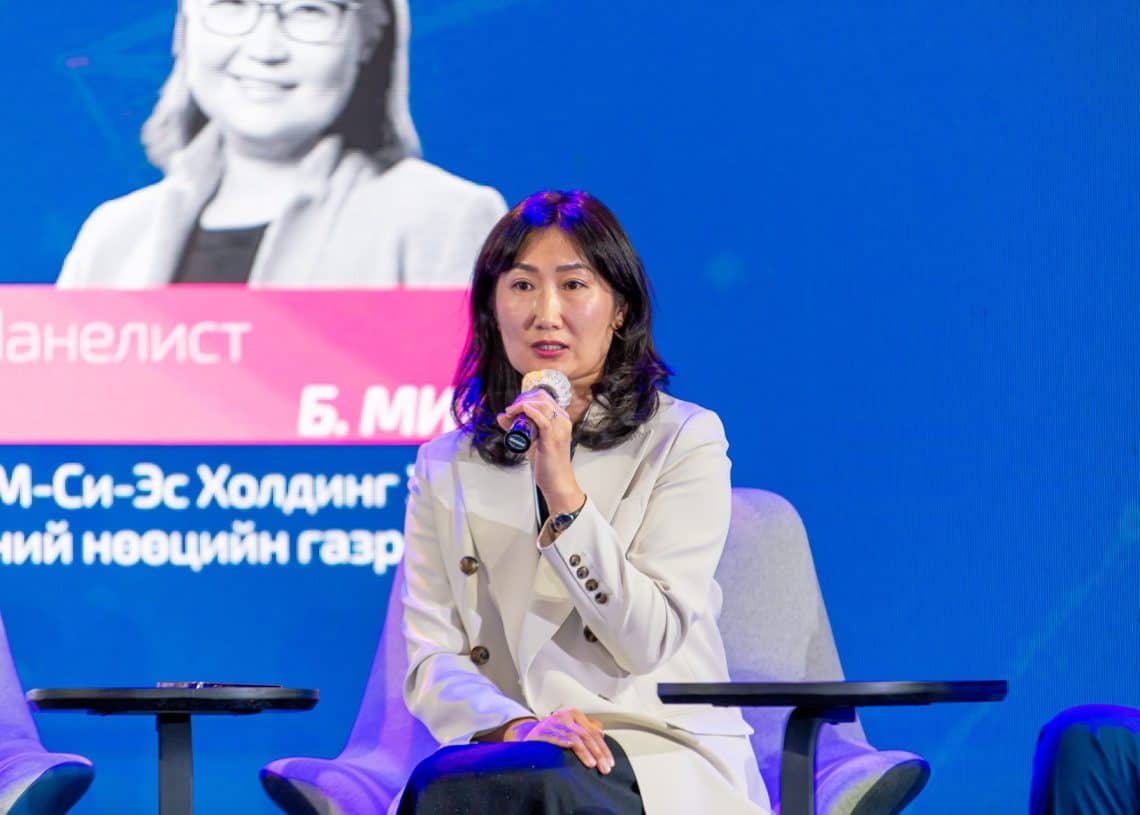 Хөнгөн пиджак өмссөн эмэгтэй "Монголын хөдөлмөрийн зах зээлийн чуулга уулзалт 2024"-ийн тайзан дээр сууж байхдаа микрофоноор ярьж байна. "панелист" зэрэг кирилл бичээс бүхий том цэнхэр дэлгэцийн өмнө тэрээр "хүний нөөц"-ийг сайжруулах "шийдэл хайсан" стратегийн талаар ярилцаж байна. . - Business.MN