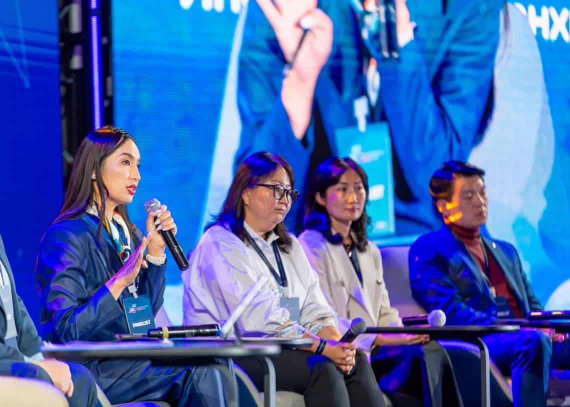 Дөрвөн хүний бүрэлдэхүүнтэй баг тайзан дээр сууж, тус бүр нь албан ёсны хувцас өмссөн байна. Тэдний ард байгаа том дэлгэц нь тэдний томруулсан дүрсийг харуулах үед нэг эмэгтэй микрофон руу ярьж байна. Энэ нь 2024 оны дээд хэмжээний уулзалт болж, Монголын хөдөлмөрийн зах зээл эсвэл хүний нөөц зэрэг сэдвүүдийг хэлэлцэх бололтой. - Business.MN