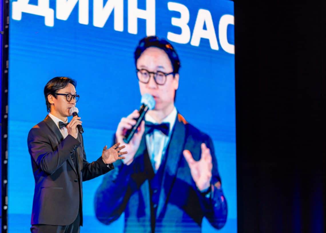 2024 оны Монголын хөдөлмөрийн зах зээлийн дээд хэмжээний уулзалтын үеэр хар өнгийн костюмтай, нүдний шил зүүсэн эр тайзан дээр микрофоноор ярьж байна. Түүний ард том дэлгэцэн дээр түүний ижил костюмтай, ярьж буй дүр төрхийг харуулсан нь үзэгчидтэй харьцаж байгааг онцолж байна. - Business.MN