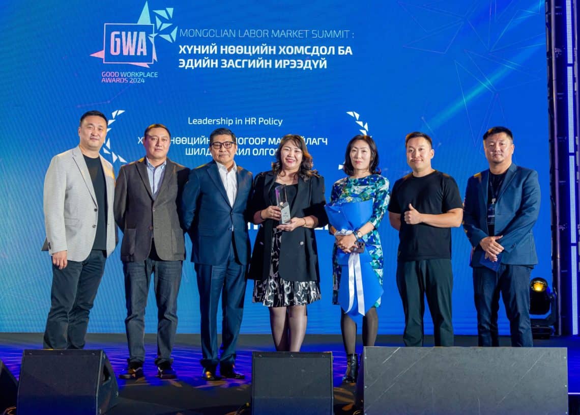 2024 оны Монголын хөдөлмөрийн зах зээлийн дээд хэмжээний уулзалтын тайзан дээр долоон хүний бүрэлдэхүүнтэй баг зогсож байна. Тэд ажил хэрэгч хувцас өмссөн бөгөөд хүний нөөцийн шилдэг хоёр шагналтай. Арын дэвсгэр нь цэнхэр өнгөтэй, үйл явдлын дэлгэрэнгүй мэдээлэл, Good Workplace Awards 2023-ын лого зэргийг агуулсан. - Business.MN