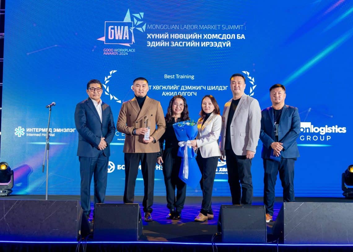 2024 оны Монголын хөдөлмөрийн зах зээлийн дээд хэмжээний уулзалтын тайзан дээр таван хүний бүрэлдэхүүнтэй баг зогсож байна. Нэг хүн цэцгийн баглаа барьдаг. Арын том дэлгэц нь хүний нөөцтэй холбоотой үйл явдлын дэлгэрэнгүй мэдээллийг харуулдаг. Тайзны гэрэлтүүлэг нь харагдахуйц болж, тайзны мэргэжлийн уур амьсгалыг сайжруулдаг. - Business.MN