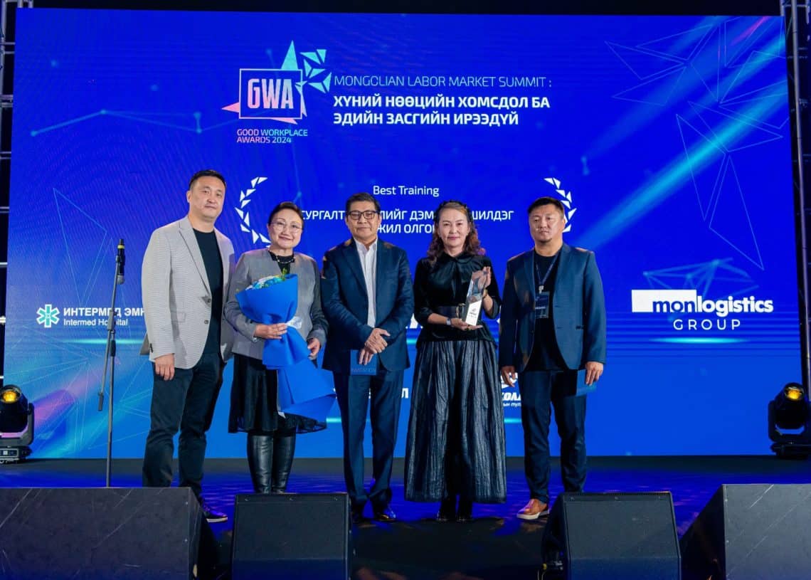 "Монголын хөдөлмөрийн зах зээлийн чуулга уулзалт-2024"-ийн тайзан дээр янз бүрийн логогоор чимэглэсэн цэнхэр цагаан дэвсгэр дээр таван хүн зогсож байна. Тайзны хурц гэрэл дунд нэг хүн баглаа цэцэг бариад гэрэл зураг авахуулж, хүний нөөцөд анхаарлаа хандуулж байгаагаа онцолж байна. - Business.MN