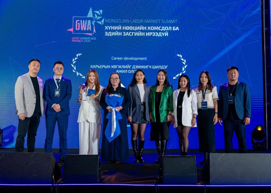 2024 оны Монголын хөдөлмөрийн зах зээлийн дээд хэмжээний уулзалтын тайзан дээр найман хүний бүрэлдэхүүнтэй баг зогсож байна. Албан ёсны хувцас өмссөн тэд тайзны гэрэлтүүлгээр гэрэлтдэг бол үйл явдлын дэлгэрэнгүй мэдээлэл бүхий цэнхэр туг нь тэдний байгаа байдлыг онцолж, хүний нөөцийн талаар ярилцах тайзыг тавьжээ. - Business.MN