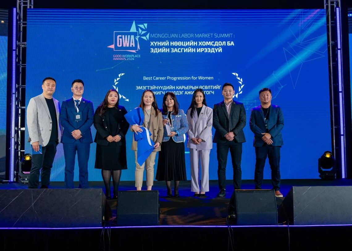 Монголын хөдөлмөрийн зах зээлийн дээд хэмжээний уулзалтын үеэр найман хүний бүрэлдэхүүнтэй хэсэг тайзан дээр зогсож байна. Төвд байгаа эмэгтэй цом барьдаг. Арын дэвсгэр дээр "Монголын хөдөлмөрийн зах зээлийн дээд хэмжээний уулзалт 2024" болон "Эмэгтэйчүүдийн карьерийн шилдэг дэвшил" гэсэн гадаад хэл дээрх бичвэрийг харуулдаг. - Business.MN