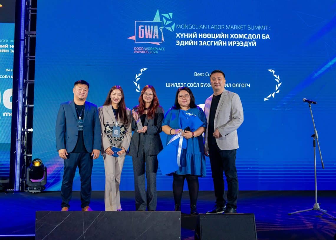 2024 оны Монголын хөдөлмөрийн зах зээлийн дээд хэмжээний уулзалтын тайзан дээр таван хүн зогсож байна. Арын арын том дэлгэц нь үйл явдалтай холбоотой текстийг харуулдаг. Тус хамтлагийнхан шагнал авсныхаа дараа баярлаж, зургаа авахуулж байгаа нь хүний нөөцийн өнөөгийн зах зээл дэх чухал ач холбогдлыг харуулсан юм. - Business.MN