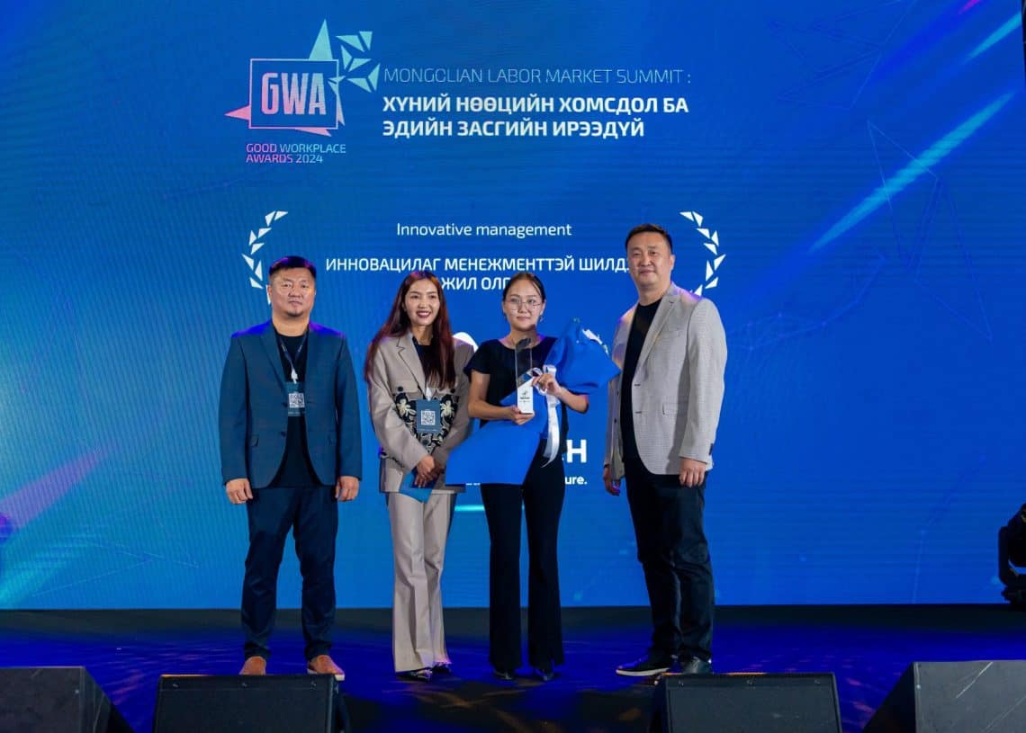 Монголын хөдөлмөрийн зах зээлийн дээд хэмжээний уулзалтын тайзан дээр дөрвөн хүн зогсож байна. Нэг хүн бахархалтайгаар шагналаа барьж байгаа бол цаана нь "Сайн ажлын байрны шагнал 2023" гэж бичсэн байна. Хүний нөөцийн мэргэжилтнүүд болох багийнхан албан ёсны хувцас өмсөн тайзны гэрэл дор инээмсэглэж байна. - Business.MN