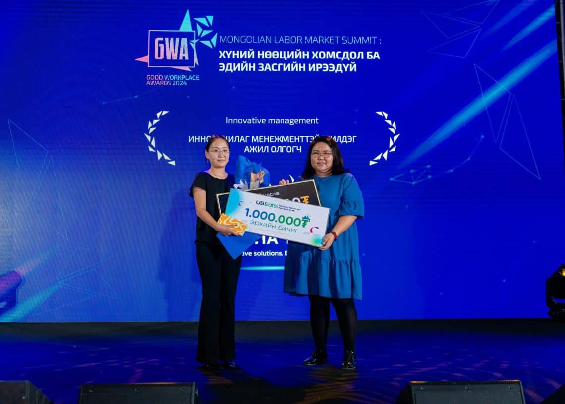 Монголын хөдөлмөрийн зах зээлийн дээд хэмжээний уулзалтын тайзан дээр хоёр эмэгтэй зогсож байна. Нэг нь "Шинэлэг менежмент"-ийн гэрчилгээ, 1,000,000₮-ийн их хэмжээний чек эзэмшиж, хүний нөөцийн мэдлэгээ үнэлдэг. Арын дэвсгэр дээр үйл явдлын гарчиг, лого харагдана. - Business.MN