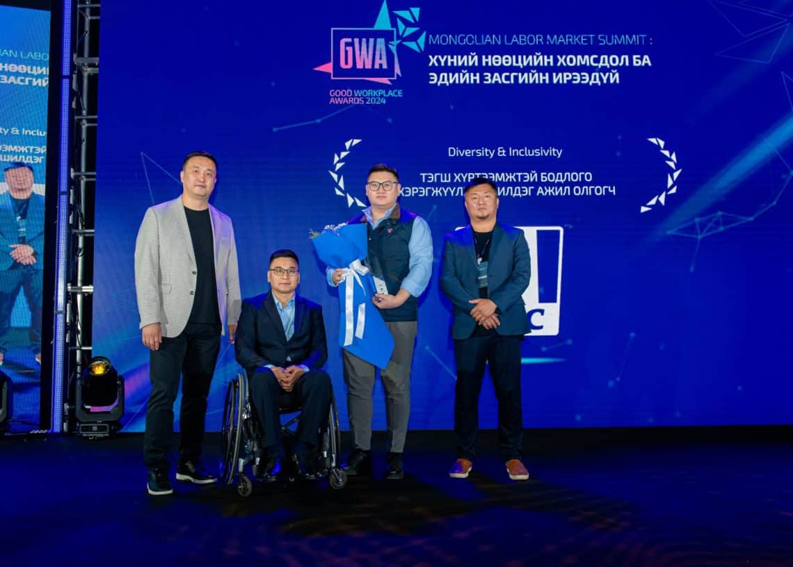 Дөрвөн хүн нэг нь тэргэнцэртэй, цэцгийн баглаа, гэрчилгээ барьчихсан тайзан дээр зогсож байна. Арын дэвсгэр нь цэнхэр өнгөтэй, 2024 оны дээд хэмжээний уулзалт, Хүний нөөцийн асуудлыг онцлон тэмдэглэсэн, Монголын Хөдөлмөрийн зах зээлийн дээд хэмжээний уулзалт, Олон янз байдал & Хамгаалалтад анхаарлаа хандуулсан бичвэрүүдээр чимэглэгдсэн байна. - Business.MN