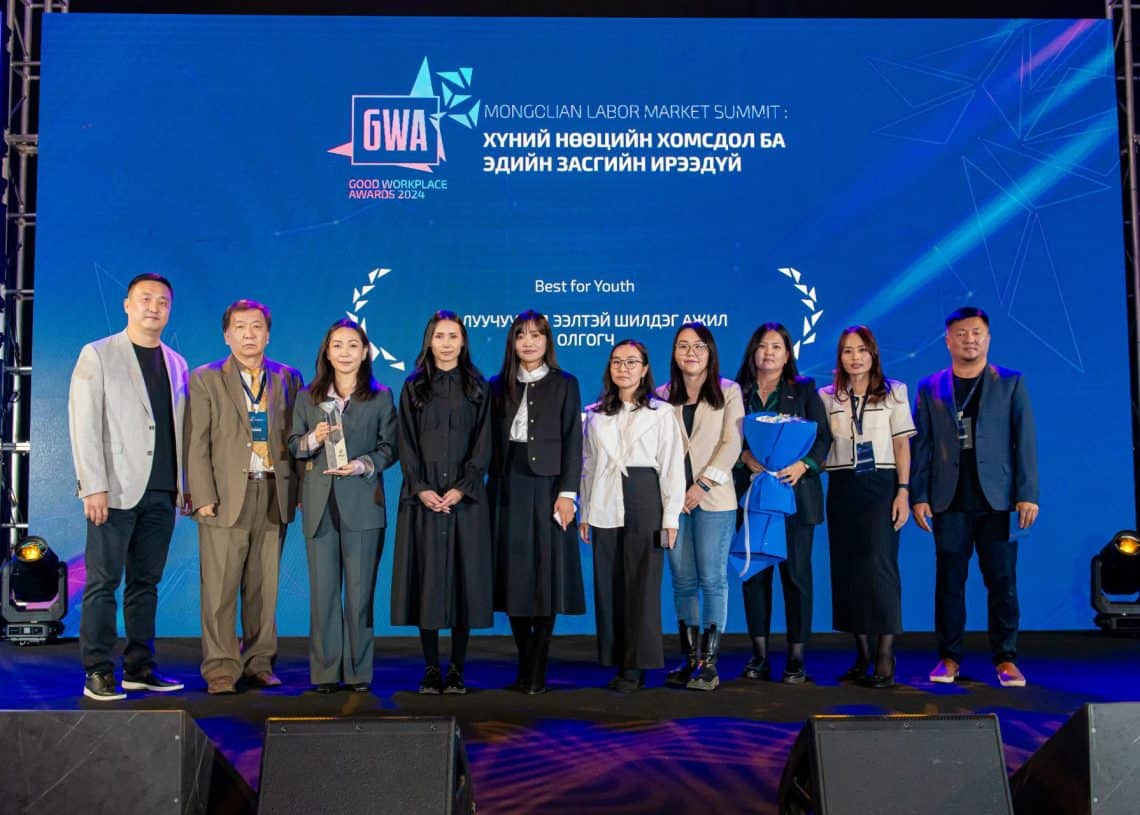 Монголын хөдөлмөрийн зах зээлийн дээд хэмжээний уулзалт 2024 дээр есөн хүний бүрэлдэхүүнтэй хэсэг тайзан дээр зогсож, инээмсэглэн гэрэл зураг авахуулж байна. Цэнхэр дэвсгэр дээр үйл явдлын лого болон бичвэрийг кирилл үсгээр харуулсан нь хүний нөөцийн санаачлагуудаас асуудлын шийдлийг олох амлалтаа онцолж байна. - Business.MN