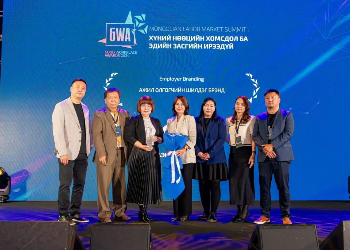 Нэг арга хэмжээнд таван эрэгтэй, хоёр эмэгтэй долоон хүний бүрэлдэхүүнтэй баг тайзан дээр зогсож байна. Тэд хүний нөөцийн дээд хэмжээний уулзалтын үеэр эрэлхийлсэн шийдлүүдийг тусгасан "Сайн ажлын байрны шагнал 2023"-ын арга хэмжээний лого, монгол бичгээр бичсэн туг, ард нь албан ёсны хувцас өмссөн байна. - Business.MN