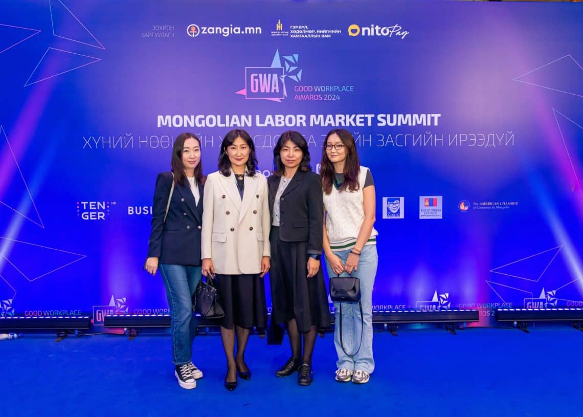 Монголын хөдөлмөрийн зах зээлийн дээд хэмжээний уулзалт 2024 дээр зэрэгцэн зогсон инээмсэглэж буй дөрвөн хүн арга хэмжээний лого, бичвэрээр чимэглэсэн цэнхэр дэвсгэрийн өмнө зургаа авахуулж байна. Өдөр тутмын болон ажил хэрэгч хувцсыг хослуулсан тус групп санаа бодлоо идэвхтэй солилцож, хүний нөөцөд өөрсдийн туршлагаа тусгадаг. - Business.MN
