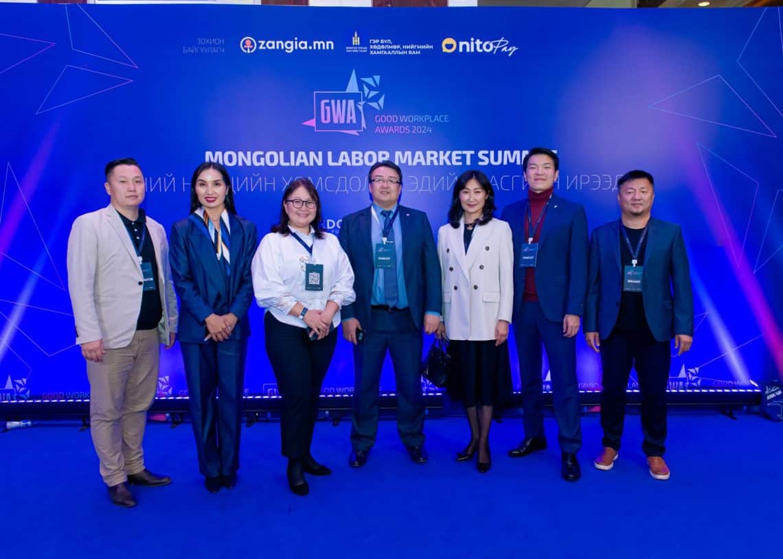 Монголын хөдөлмөрийн зах зээлийн чуулга уулзалт 2024 арга хэмжээний цэнхэр дэвсгэрийн өмнө долоон хүний бүрэлдэхүүнтэй хэсэг хүмүүс ажил хэрэгч хувцастай, тус бүрдээ нэрийн тэмдэг зүүсэн байна. Энэхүү дээд хэмжээний уулзалт нь хүний нөөцийн тулгамдсан асуудлуудыг шийдвэрлэхийг зорьж байгаа бөгөөд арын дэвсгэр нь лого, үйл явдлын графикийг агуулсан болно. - Business.MN