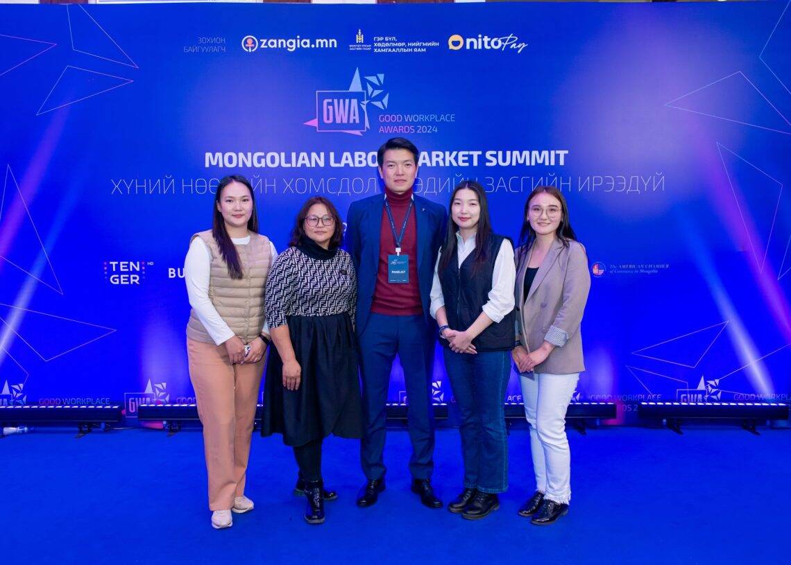 Монголын хөдөлмөрийн зах зээлийн дээд хэмжээний уулзалтад таван хүний бүрэлдэхүүнтэй баг цэнхэр хивсэн дээр зогсож байна. Арын дэвсгэр дээр арга хэмжээний лого болон "Сайн ажлын байрны шагнал 2023" гэсэн бичвэрийг харуулсан байна. Тэд хүний нөөцийн асуудлын санаачилгын онцлох үйл явдлуудыг тэмдэглэж, гэрэл зураг авахаар инээмсэглэж байна. - Business.MN