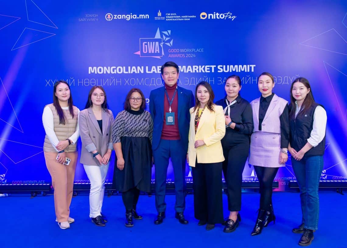 Монголын хөдөлмөрийн зах зээлийн дээд хэмжээний уулзалт 2024 дээр тайзан дээр инээмсэглэж буй найман хүний бүрэлдэхүүнтэй баг хамт олон. Ажил хэрэгч энгийн хувцас өмссөн тэд арга хэмжээний лого, нарийн ширийн зүйлсээр чимэглэгдсэн цэнхэр дэвсгэрийн өмнө зургаа авахуулж, хүний нөөцийн асуудалд хамтран ажиллах сэтгэлийг шингээжээ. - Business.MN