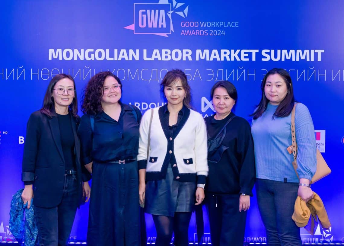 "Монголын хөдөлмөрийн зах зээлийн дээд хэмжээний уулзалт", "Сайн ажлын байрны шагнал 2024" гэсэн бичиг бүхий цэнхэр дэвсгэрийн өмнө таван хүн хамтдаа инээмсэглэн зогсож байна. Тэд хагас албан ёсны хувцас өмсөж, ирэх жилийн хүний нөөцийн (хүний нөөц) ололт амжилтаа тэмдэглэж байна.
- Business.MN