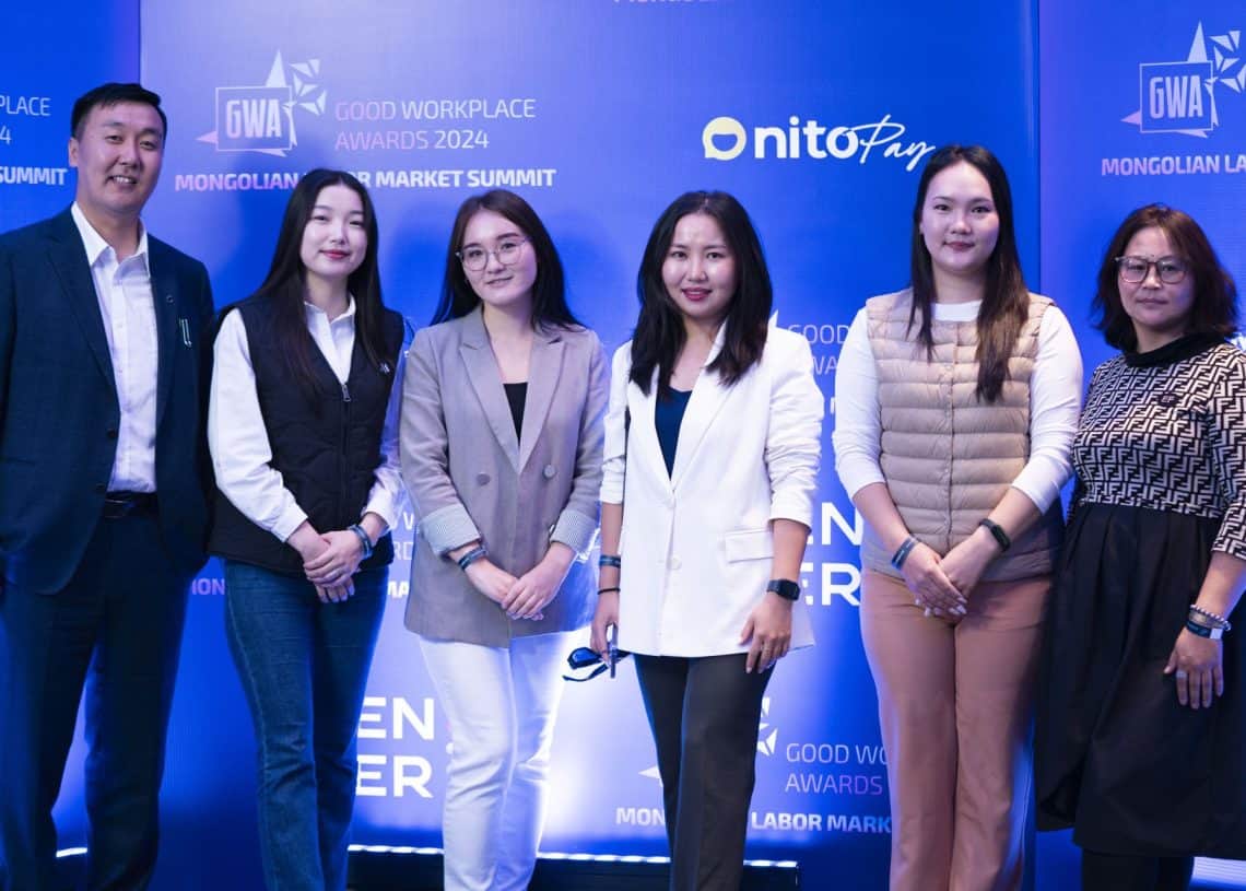Зургаан хүний бүрэлдэхүүнтэй хэсэг хүмүүс "Good Workplace Awards 2024" болон "Монголын хөдөлмөрийн зах зээлийн дээд хэмжээний уулзалт 2024"-ын лого бүхий цэнхэр дэвсгэрийн өмнө зогсож байна. Тэд ажил хэрэгч энгийн хувцас өмсөж, камер руу инээмсэглэж байгаа нь янз бүрийн шийдэл хайсан гэдгээ илэрхийлж байна. хүний нөөцийн сорилтууд. - Business.MN