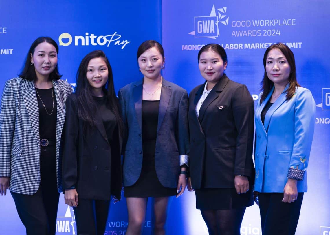 Асуудал, шийдвэрлэх арга замыг хэлэлцдэг Монголын хөдөлмөрийн зах зээлийн чуулга уулзалт 2024 арга хэмжээнд албан ёсны хувцастай таван хүн хамтдаа зогсож байна. Цэнхэр дэвсгэр дээр хүний нөөцийн менежментийн шинэчлэлийг онцлон харуулсан "Сайн ажлын байрны шагнал 2024", "Онитопай" лого харагдаж байна. - Business.MN