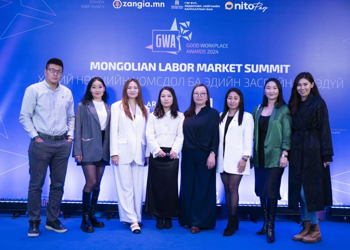 2024 оны Монголын хөдөлмөрийн зах зээлийн дээд хэмжээний уулзалтын цэнхэр дэвсгэрийн өмнө найман хүний бүрэлдэхүүнтэй хэсэг зогсож байна. Тэд ажил хэрэгч, ухаалаг энгийн хувцас өмсөж, халуун дулаан инээмсэглэж, хүний нөөцийг бахархалтайгаар төлөөлдөг. - Business.MN