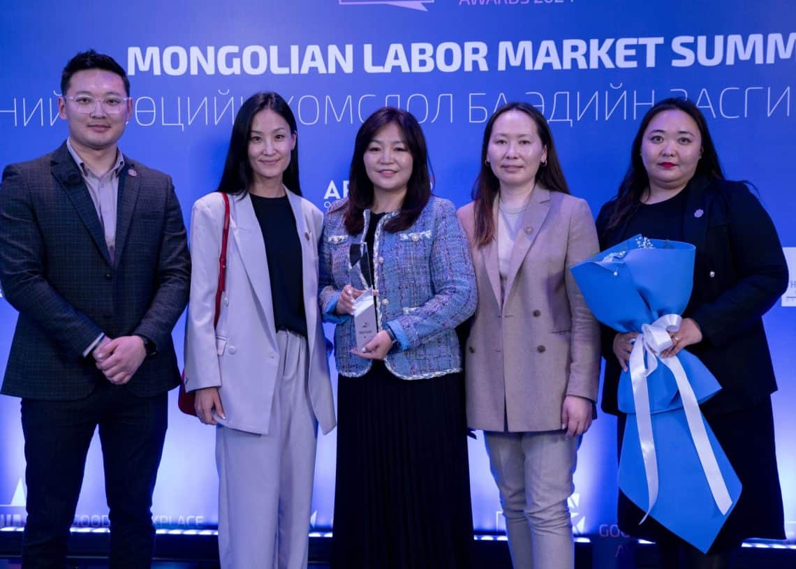 "Монголын хөдөлмөрийн зах зээлийн чуулга уулзалт"-ын тайзны өмнө таван хүний бүрэлдэхүүнтэй баг зогсож байна. Төвийн хүн цом бардам барьдаг бол баруун талд байгаа хоёр нь цэнхэр, цагаан цэцгийн баглаа бэлэглэж байна. Эргэн тойрон инээмсэглэлээр тэд багаар ажиллах, хүний нөөцөд шийдэл гаргах амлалтыг илэрхийлдэг. - Business.MN