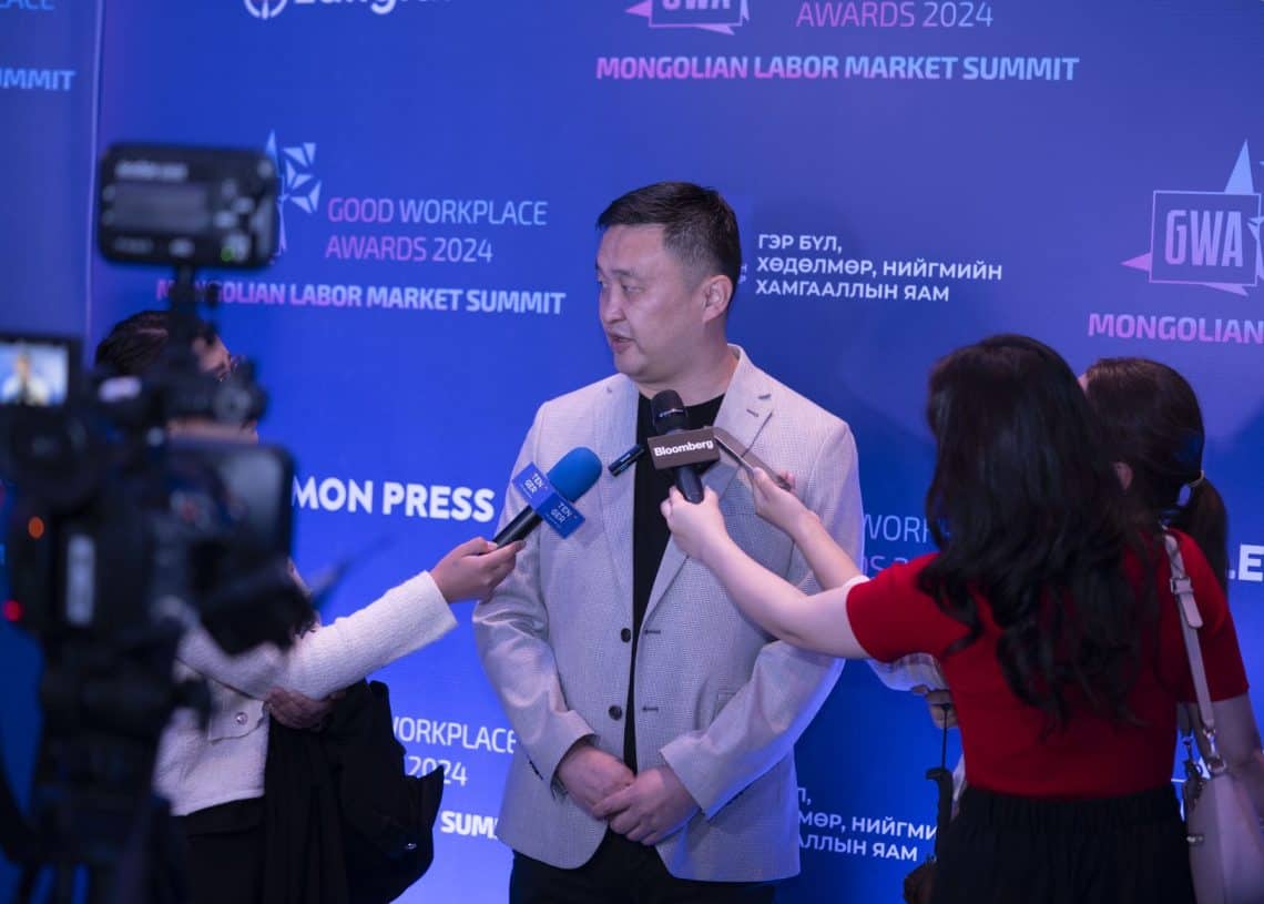 Цайвар саарал костюмтай эр Монголын хөдөлмөрийн зах зээлийн дээд хэмжээний уулзалт 2024 дээр сэтгүүлчидтэй ярилцаж байна. Лого болон үйл явдлын нэрээр чимэглэсэн арын дэвсгэр нь камерын багийнхны авсан динамик солилцоог дүрсэлжээ. - Business.MN