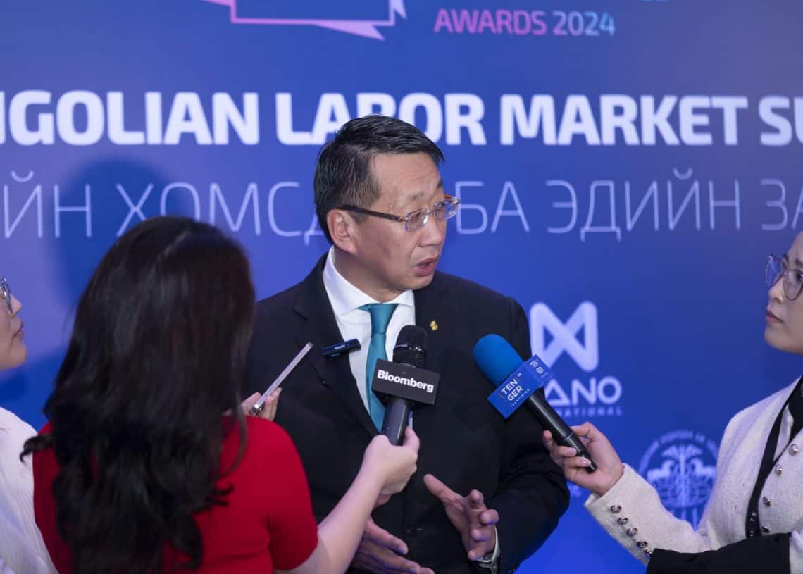 Микрофон барьсан гурван сэтгүүлч ярилцлага өгч байхад костюмтай эр ярьж байна. Арын дэвсгэр дээр хүний нөөц, шагналын талаарх хэлэлцүүлгийг онцолж, Монголын хөдөлмөрийн зах зээлийн чуулга уулзалт 2024-тэй холбоотой баннеруудыг харуулсан байна. - Business.MN