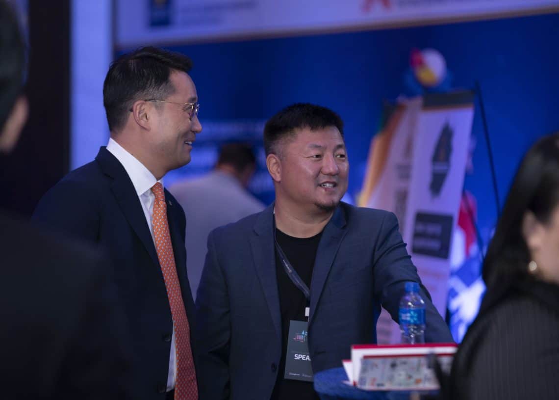 Албаны хувцастай хоёр хүн танхимын арга хэмжээний үеэр харилцан яриа өрнүүлж байна. Нэг нь костюм, зангиа, нөгөө нь зангиагүй хослол өмсдөг. Тэд товхимол, усны савтай ширээний дэргэд цэнхэр гэрэлтүүлэгтэй зогсож байна. 2024 оны дээд хэмжээний уулзалтад оролцогчид болон баннерууд цаана байгаа. - Business.MN