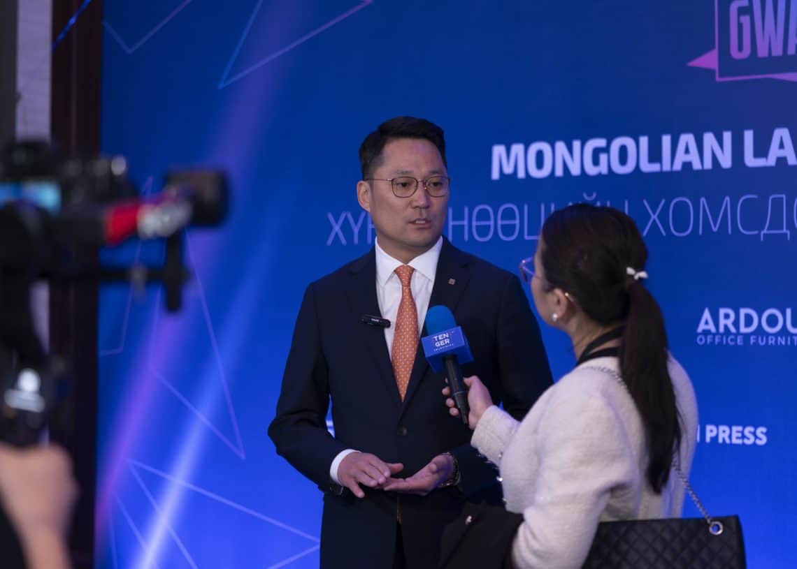 2024 оны Монголын хөдөлмөрийн зах зээлийн дээд хэмжээний уулзалтын үеэр гэзэгтэй эмэгтэйн барьж буй микрофонд костюмтай эр ярьж байна. Хүний нөөц, салбарын чиг хандлагын талаарх ойлголтыг агуулсан камер, лого нь ард харагдаж байна. - Business.MN