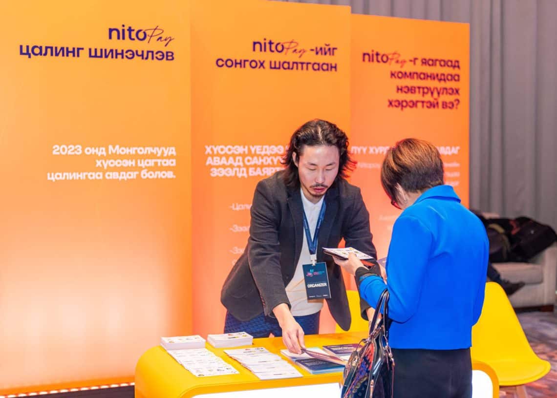 Монголын хөдөлмөрийн зах зээлийн тухай сурталчилгааны материал бүхий ширээний ард зогсож буй эрэгтэй утас барьсан эмэгтэйтэй харьцаж байна. Арын дэвсгэр дээр Монгол бичвэр, Nito брэндийн лого бүхий улбар шар өнгийн хавтангууд нь Summit 2024-ийн хэлэлцүүлгийг илтгэж байна. - Business.MN