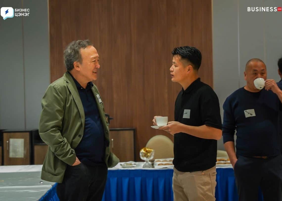 Two men are having a conversation while holding cups in a room with a large table and refreshments in the background. Another man is drinking from a cup behind them. All are wearing casual business attire.