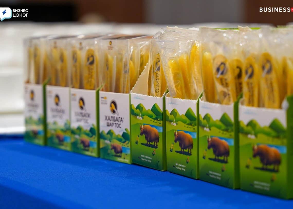 Boxes of packaged cheese sticks are lined up on a blue tablecloth. The packaging features illustrations of a cow grazing in green fields with mountains in the background.