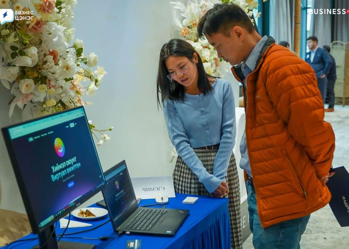 A woman and a man stand at a display table with a laptop and brochures on a blue tablecloth. The screen shows a colorful graphic. Floral decorations are in the background. They appear to be at a business event.