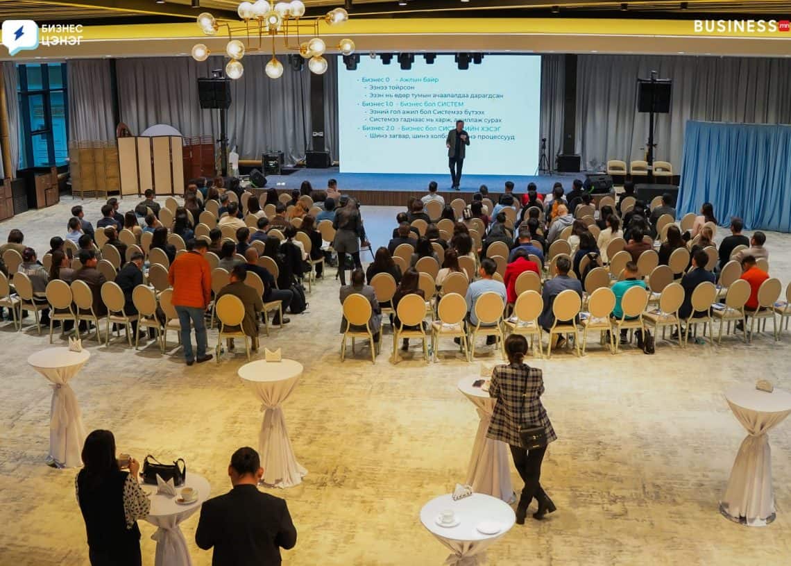 A speaker addresses an audience seated in a spacious event hall. The room features round tables in the foreground and a screen displaying a presentation behind the speaker. The attendees are focused on the presentation.
