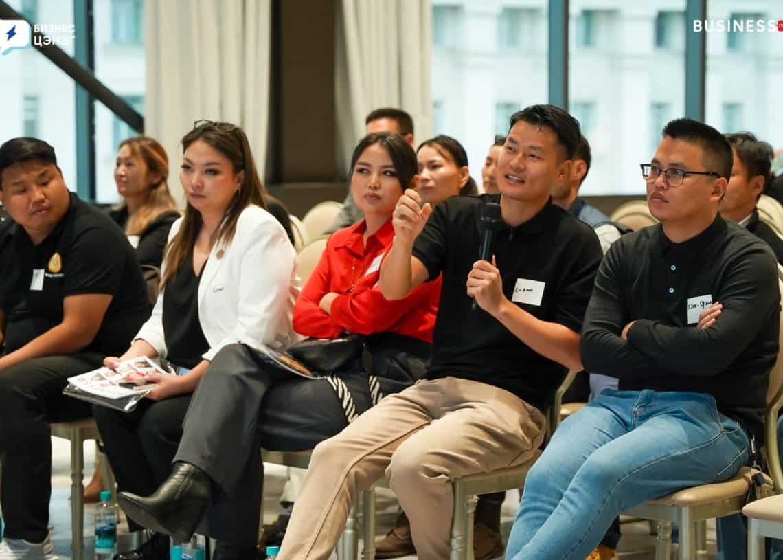 A group of people sitting in chairs at an indoor event. Several individuals are listening attentively, with one person speaking into a microphone. They are dressed in casual and professional attire.
