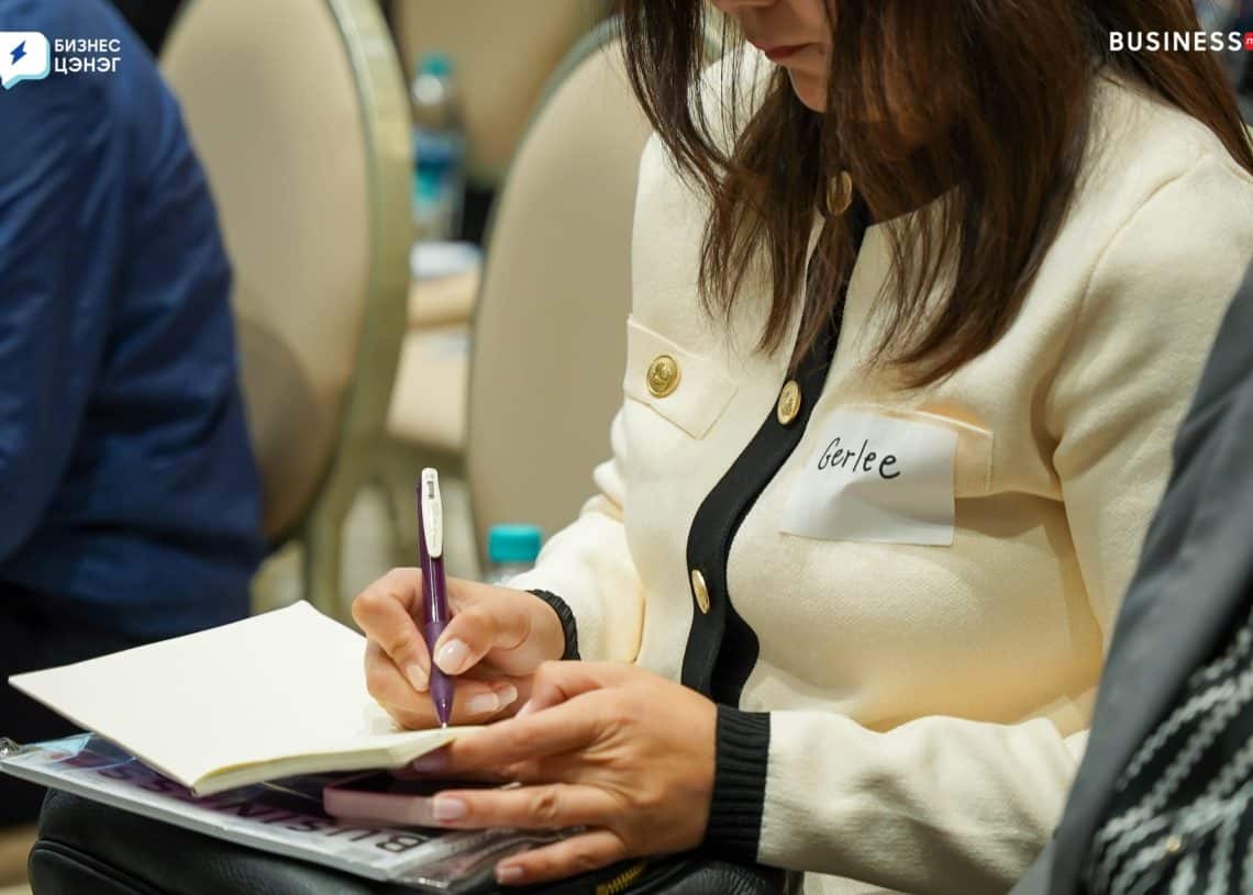 A woman with long brown hair is taking notes in a notebook during a conference. She is wearing a white and black cardigan with gold buttons. A name tag is visible on her chest. Other attendees are seated nearby.