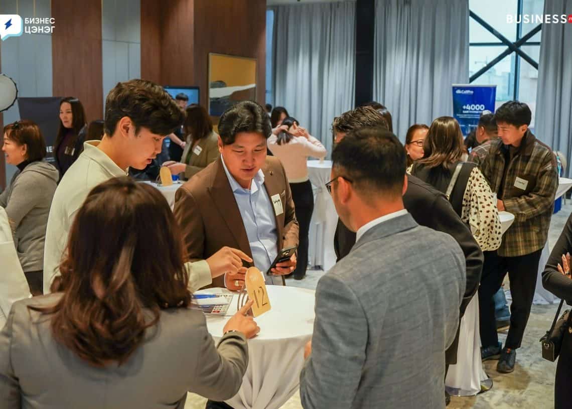 A group of people in formal attire engage in discussions around a conference table. They appear to be networking at a professional event, with name tags visible. 력, 식들, and other words can be seen in the background signage.