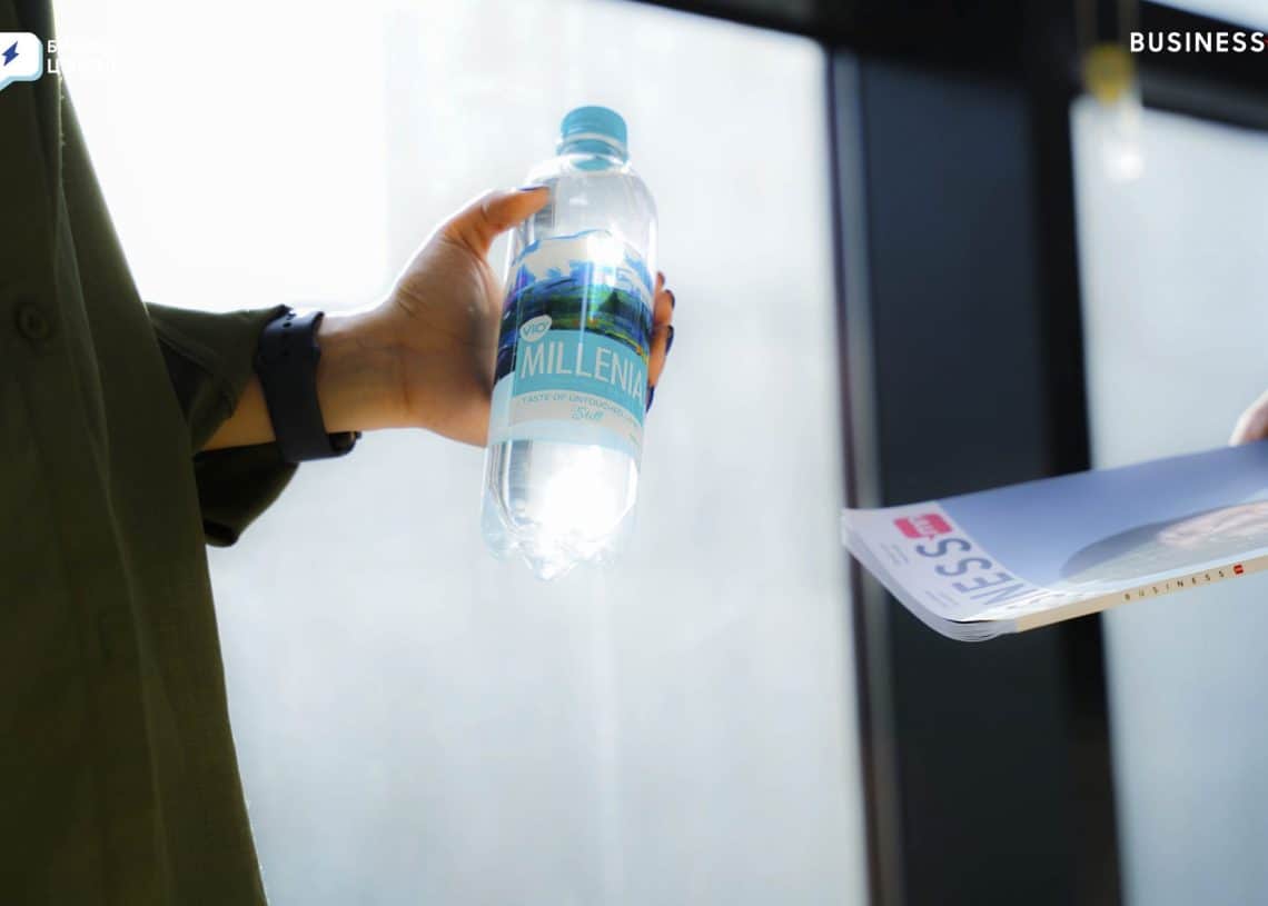 A person holds a bottle of water in their right hand, labeled "Millennia," while handing a business magazine to someone else indoors. The background is softly lit, with large windows.